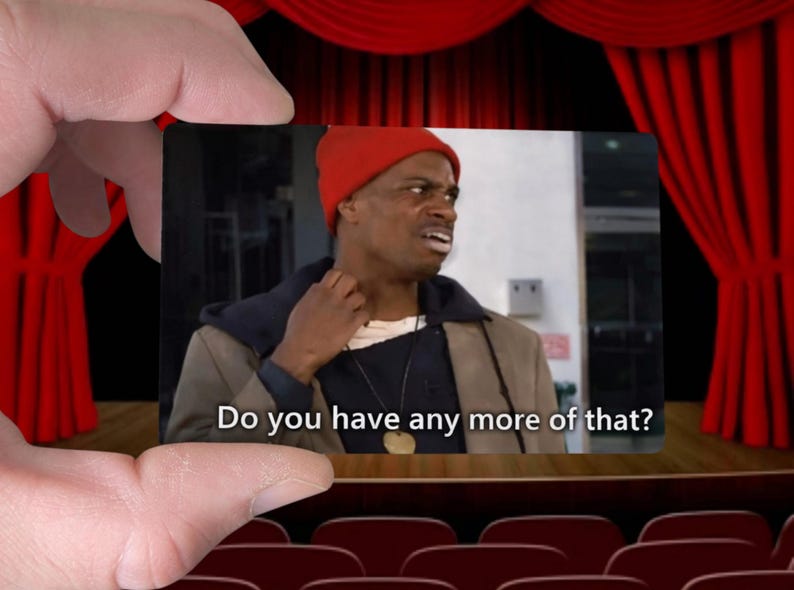 May include: A meme featuring a man in a red beanie and a brown jacket, with the text "Do you have any more of that?" The image is held by a hand against a backdrop of a theater stage with red curtains.