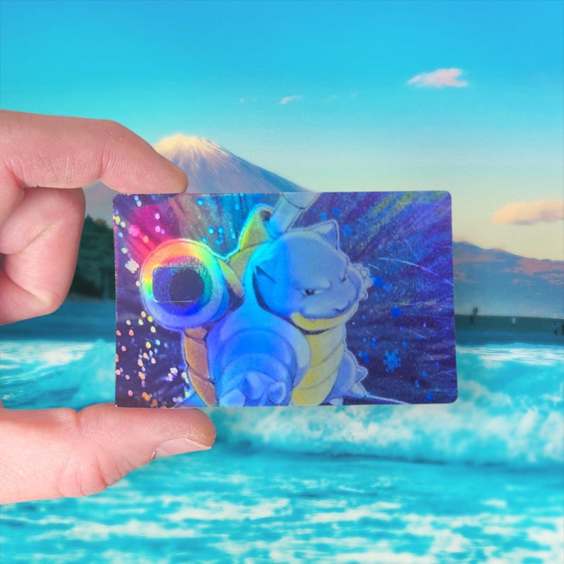 May include: A holographic trading card featuring a blue cartoon character with a shell and water cannons. The card is held between two fingers, with a scenic background of a mountain and ocean.