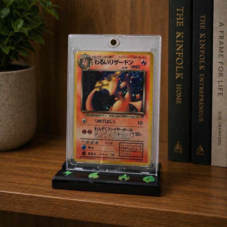 Puede incluir: Una tarjeta Pok&eacute;mon Charizard enmarcada en una funda protectora, exhibida en una estanter&iacute;a de madera. La tarjeta presenta texto japon&eacute;s y una ilustraci&oacute;n en naranja y amarillo fuego. Hay libros en el fondo.