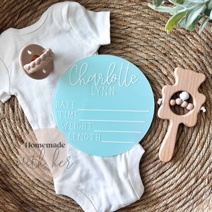 May include: A light blue round baby announcement sign with the name Charlotte Lynn and spaces to fill in the date, time, weight, and length. The sign is on a woven basket with a white onesie and a wooden baby rattle.