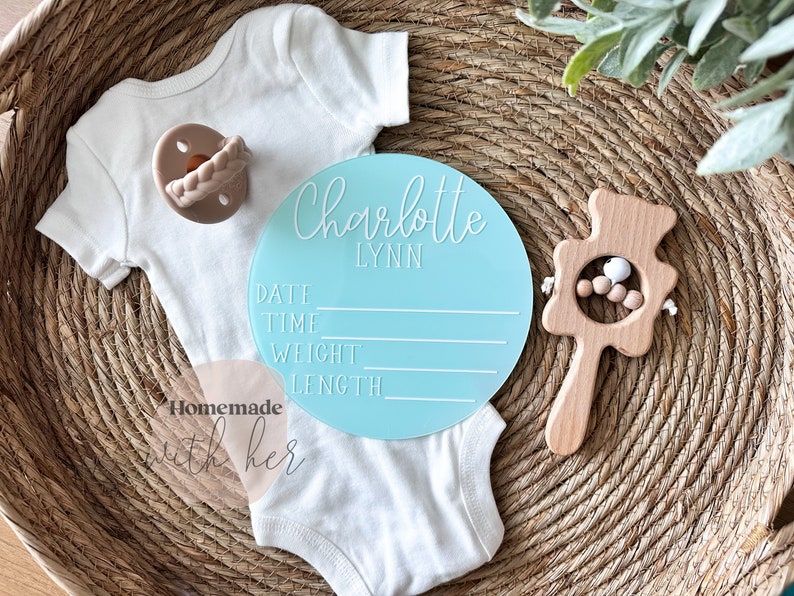 May include: A light blue round baby announcement sign with the name Charlotte Lynn and spaces to fill in the date, time, weight, and length. The sign is on a woven basket with a white onesie and a wooden baby rattle.