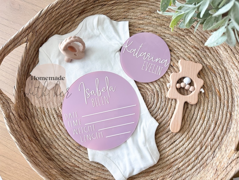 May include: Two pink acrylic baby announcement plaques. One plaque says "Isabela Belen" and has lines for date, time, weight, and length. The other plaque says "Katarina Evelin".