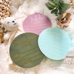 May include: Three round wooden plaques with white lettering. The plaques are green, pink, and blue. Each plaque has a name, "Sebastian James", "Isabela Belen", and "Charlotte Lynn", and spaces to record the date, time, weight, and length. The text "Homemade with her" is visible in the bottom right corner.