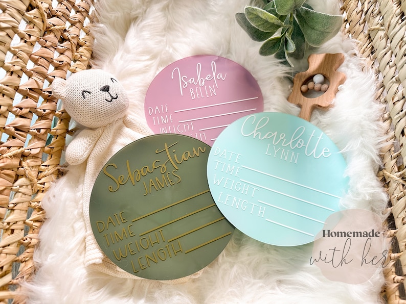 May include: Three round wooden plaques with white lettering. The plaques are green, pink, and blue. Each plaque has a name, "Sebastian James", "Isabela Belen", and "Charlotte Lynn", and spaces to record the date, time, weight, and length. The text "Homemade with her" is visible in the bottom right corner.