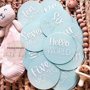 May include: A set of ten turquoise blue baby milestone discs with white text. The discs feature the phrases "Hello World", "One Month", "Two Months", "Three Months", "Four Months", "Five Months", "Six Months", "Seven Months", "Eight Months", and "Ten Months".
