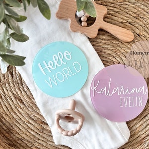 May include: Two round acrylic baby announcement signs. One is light blue with the text "Hello World" in white. The other is light purple with the text "Katarina Evelin" in white.