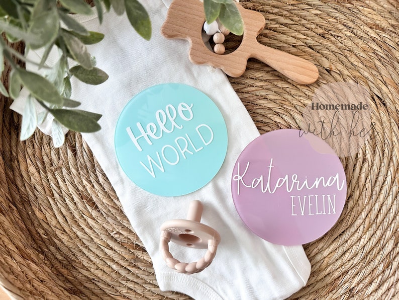 May include: Two round acrylic baby announcement signs. One is light blue with the text "Hello World" in white. The other is light purple with the text "Katarina Evelin" in white.