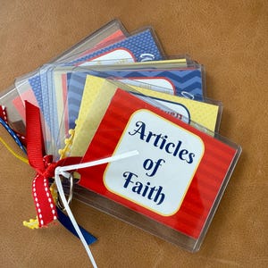 May include: A collection of laminated cards, bound together with a metal ring and colorful ribbons. The front card reads "Articles of Faith" in a white, stylized font against a red and white background. The other cards have various patterns and colors.
