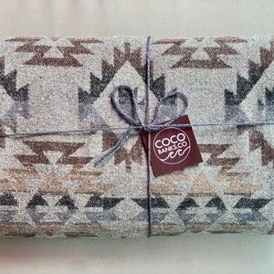 Puede incluir: Una manta doblada con un patrón geométrico en tonos grises, marrones y beige. La manta está atada con hilo gris y una etiqueta de COCO BANKS. El diseño de la manta presenta formas triangulares y en zigzag repetidas.