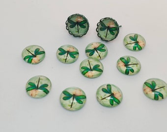 May include: A collection of dragonfly-themed jewelry and decorative cabochons. The earrings have a dark green filigree setting. The cabochons and earrings feature a dragonfly design with green wings and a light green background with a floral pattern.