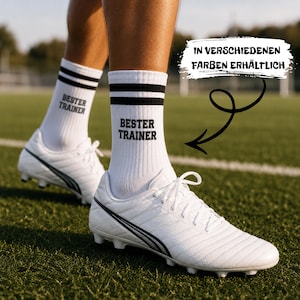 May include: White crew socks featuring two black horizontal stripes at the top and the black text 'BESTER TRAINER' printed on the side. The socks are worn with white football boots. A graphic element with the text 'IN VERSCHIEDENEN FARBEN ERHÄLTLICH' points to the socks.