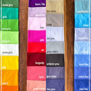 Puede incluir: Una carta de colores que muestra cuarenta y dos muestras de tela para camisetas, dispuestas en cuatro columnas verticales. El texto T-SHIRT FARBEN es visible en la parte superior. Las muestras presentan un amplio espectro de colores, incluyendo verdes, morados, rosas, rojos, naranjas, blancos, beiges, grises, azules y negros, cada uno etiquetado con su nombre de color espec&iacute;fico.