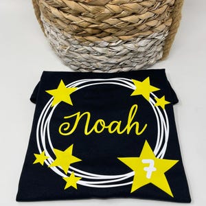 Puede incluir: Una camiseta negra con un dise&ntilde;o de corona circular blanca, estrellas amarillas y el nombre Noah en letra cursiva amarilla. Una estrella amarilla grande en la parte inferior derecha contiene el n&uacute;mero 7 en blanco. El dise&ntilde;o est&aacute; centrado.