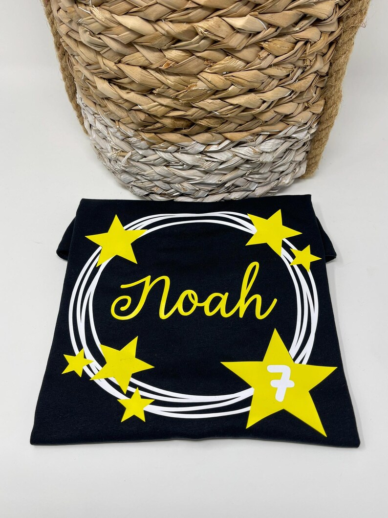 Puede incluir: Una camiseta negra con un dise&ntilde;o de corona circular blanca, estrellas amarillas y el nombre Noah en letra cursiva amarilla. Una estrella amarilla grande en la parte inferior derecha contiene el n&uacute;mero 7 en blanco. El dise&ntilde;o est&aacute; centrado.