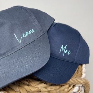 Peut inclure: Deux casquettes de baseball, une gris anthracite et une bleu marine, avec du texte cursif bleu clair. La casquette anthracite affiche le nom Leano, tandis que la casquette bleu marine affiche le nom Mac, brodés sur les panneaux avant.