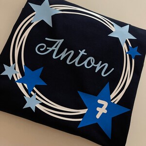 Puede incluir: Una camiseta azul marino oscuro con un dise&ntilde;o circular de l&iacute;neas blancas conc&eacute;ntricas. El nombre "Anton" est&aacute; impreso en una fuente cursiva de color azul claro en el centro. El dise&ntilde;o est&aacute; acentuado con varias estrellas de color azul claro y oscuro, con el n&uacute;mero 7 en blanco sobre una de las estrellas azul oscuro m&aacute;s grandes.
