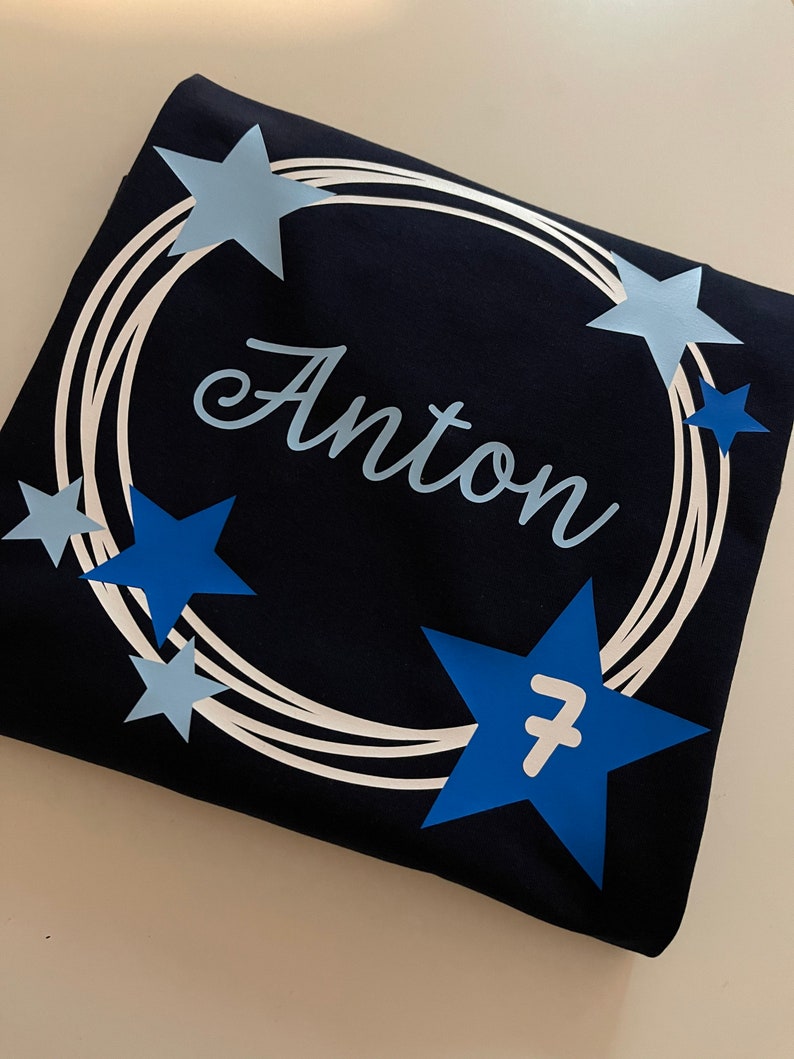 Puede incluir: Una camiseta azul marino oscuro con un dise&ntilde;o circular de l&iacute;neas blancas conc&eacute;ntricas. El nombre "Anton" est&aacute; impreso en una fuente cursiva de color azul claro en el centro. El dise&ntilde;o est&aacute; acentuado con varias estrellas de color azul claro y oscuro, con el n&uacute;mero 7 en blanco sobre una de las estrellas azul oscuro m&aacute;s grandes.