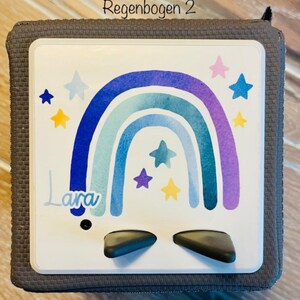 May include: A gray cube with a white face featuring a watercolor rainbow design in shades of blue and purple, with star accents. The name "Lara" is written in cursive. The text "Regenbogen 2" is at the top.