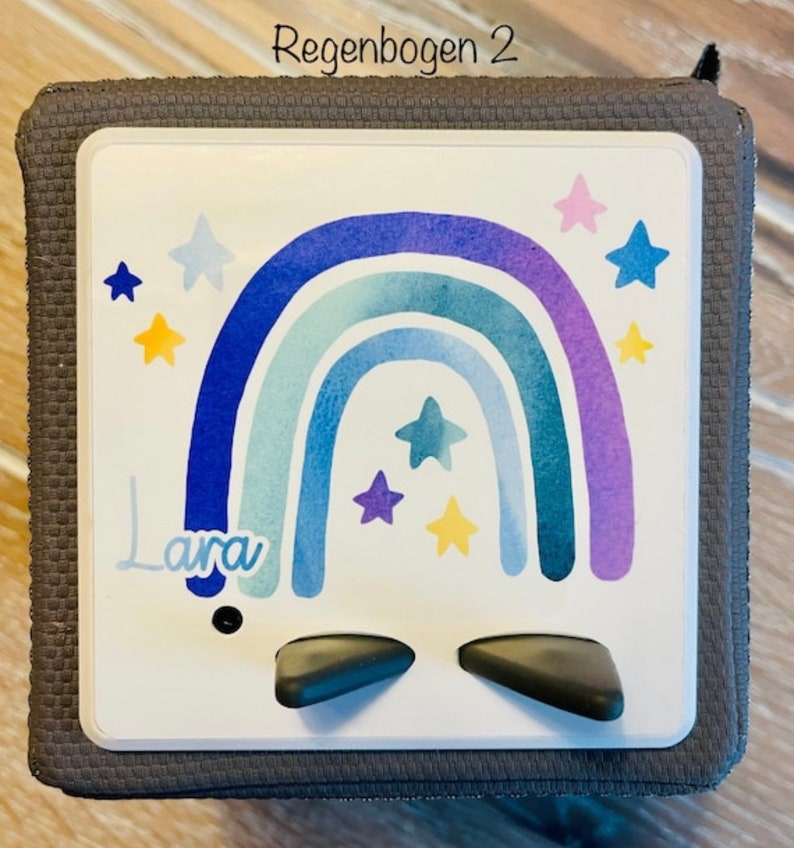 May include: A gray cube with a white face featuring a watercolor rainbow design in shades of blue and purple, with star accents. The name "Lara" is written in cursive. The text "Regenbogen 2" is at the top.