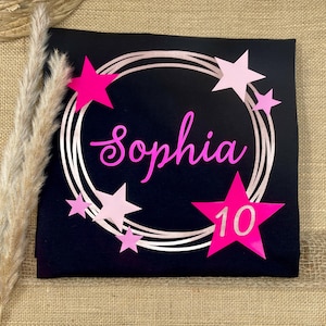 Puede incluir: Una camiseta negra con un dise&ntilde;o de marco circular en oro rosa y el nombre Sophia en letra cursiva rosa brillante. El gr&aacute;fico incluye varias estrellas en diferentes tonos de rosa y una estrella grande de color rosa brillante con el n&uacute;mero 10 en blanco.