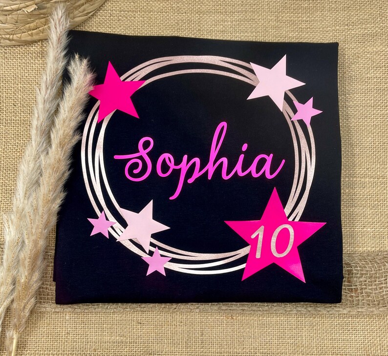 Puede incluir: Una camiseta negra con un dise&ntilde;o de marco circular en oro rosa y el nombre Sophia en letra cursiva rosa brillante. El gr&aacute;fico incluye varias estrellas en diferentes tonos de rosa y una estrella grande de color rosa brillante con el n&uacute;mero 10 en blanco.