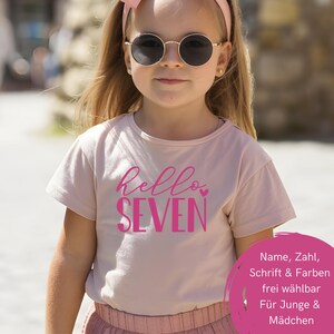 Pode incluir: Uma t-shirt de manga curta rosa claro com o texto "hello SEVEN" em letras cursivas e de imprensa rosa escuro, com um pequeno coração. A peça é usada com uma fita rosa e óculos redondos, e inclui um elemento gráfico circular.