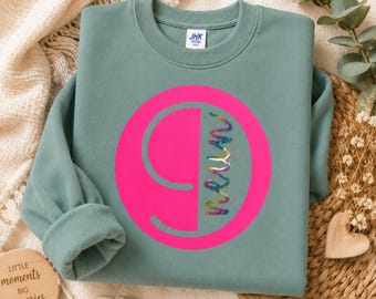 Könnte beinhalten: Ein salbeigrüner Rundhals-Pullover mit einem großen, leuchtend pinken Kreis auf der Brust. Im Inneren des Kreises befindet sich die Zahl 9 in Pink, ergänzt durch ein vertikales, mehrfarbiges, glitzerndes und strukturiertes Liniendesign.