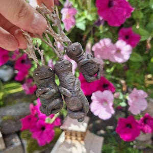 May include: Three small, hand-crafted, green and brown clay figures with twig-like appendages. The figures have rounded, bulbous heads and are hanging from a string.