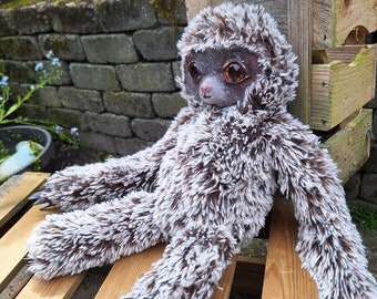 May include: A soft toy sloth with brown and white shaggy fur. The sloth has a detailed face with large, expressive eyes and claws. It is positioned on a wooden surface, with a natural background.