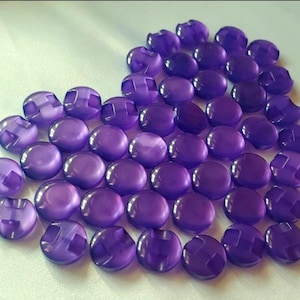 May include: A collection of round, translucent purple buttons arranged in a heart shape. Each button features a smooth, glossy surface with two holes in the center for attachment. The buttons are uniform in size and color, creating a vibrant display.