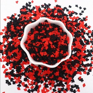 May include: A white bowl filled with red and black playing card suit confetti. The confetti includes hearts, diamonds, clubs, and spades. The confetti is scattered around the bowl on a white surface.