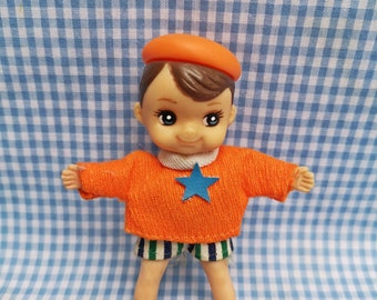 Pode incluir: Uma pequena boneca de plástico vestindo um suéter laranja, shorts listrados azul e verde e um boné laranja. A boneca tem cabelo castanho e olhos azuis.