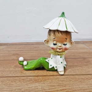 以下が含まれることがあります： 白い花形の帽子をかぶり、緑色の水玉模様が入った白い長袖シャツと、足元に白いポンポンが付いた緑色のズボンを履いた、横たわるエルフの陶器製フィギュア。首元には金色のグリッターがあしらわれた襟が付いています。