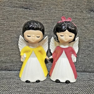 Könnte beinhalten: Zwei Keramik-Engelsfiguren mit geschlossenen Augen und schwarzen Haaren. Ein Engel trägt ein gelbes Gewand, der andere ein rotes Gewand. Beide haben weiße Flügel und halten ein kleines weißes Buch. Die Figuren stehen auf einer grau strukturierten Oberfläche.