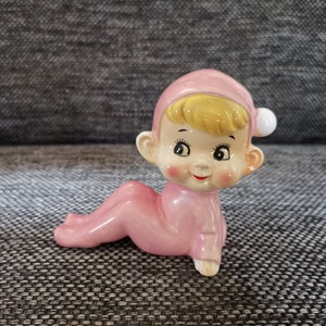 Puede incluir: Figura de cerámica de un bebé con un mono rosa y gorro a juego con un pompón blanco. El bebé está gateando, con pelo rubio, mejillas rosadas y una expresión sonriente. La figura está sobre una superficie gris texturizada.