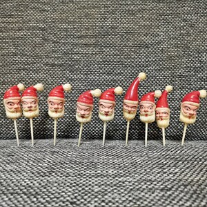 May include: Ten red and white Santa Claus head cake toppers with white sticks. The Santa heads are all facing forward with different expressions.
