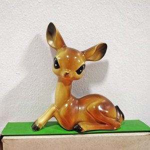 Op de afbeelding: Een vintage keramieken hertenbeeldje in rusthouding. Het hert heeft een glanzende afwerking met bruin- en beigetinten, zwarte hoeven en grote, donkere ogen. De oren zijn donkerbruin gekleurd. Het beeldje rust op een groene ondergrond.