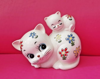 May include: White ceramic cat figurine with a smaller cat figurine on its back. Both cats have painted-on eyes and smiling mouths. The larger cat has colourful flowers painted on its body.