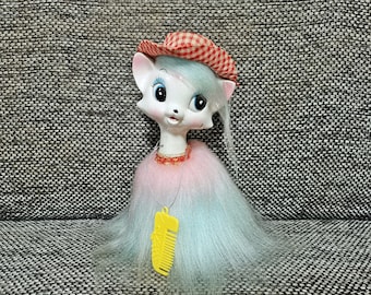 May include: A white porcelain doll with blue hair and a red and white checked hat. The doll is wearing a pink and white fur coat and a yellow plastic comb necklace.