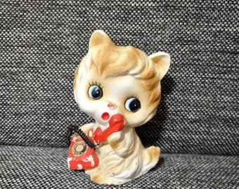 May include: A white ceramic figurine of a cat holding a red rotary phone with a black cord. The cat has brown fur and blue eyes.