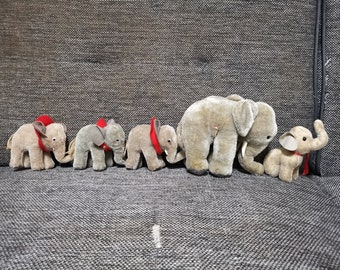 Pode incluir: Uma linha de cinco elefantes de pelúcia cinza com detalhes em tecido vermelho. Os elefantes estão dispostos em fila, com o elefante maior na parte de trás e o menor na frente.
