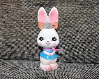 May include: A white ceramic bunny figurine with pink ears and a blue dress. The bunny has large, black eyes and a pink nose. It is wearing a pink bow with a blue flower on top.