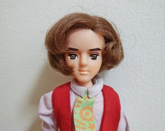 Pode incluir: Uma boneca com cabelo castanho, vestindo um colete vermelho sobre uma camisa roxa clara e calças verde claro. A boneca tem uma gravata verde com padrões florais laranja. A pele da boneca é clara e os olhos são escuros.