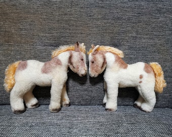 May include: Two plush toy horses are positioned facing each other. The horses have white bodies with brown spots and manes. Their tails are a light yellow color. The toys are set against a gray textured background.