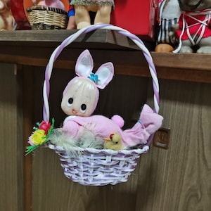 Könnte beinhalten: Eine rosa Hasenpuppe sitzt in einem lila und weißen Weidenkorb. Der Korb hängt an einem lila und weißen gedrehten Seil. Der Hase hat eine blaue Schleife auf dem Kopf und ein gelbes Küken sitzt mit ihm im Korb.