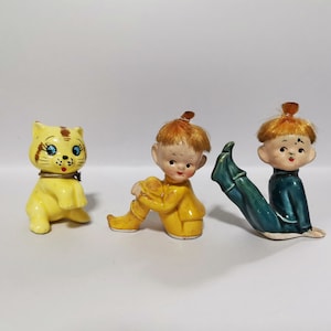 Könnte beinhalten: Drei Vintage-Keramikfiguren: eine gelbe Katze mit braunen Streifen und blauen Augen sowie zwei Figuren von Kindern mit orangefarbenem Haar. Ein Kind trägt ein gelbes Outfit, das andere ein türkisfarbenes Outfit.