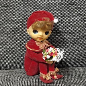 Peut inclure: Une poupée vintage vêtue d'une tenue en velours rouge, comprenant un chapeau avec un pompon blanc. La poupée tient un petit bouquet de fleurs avec une bordure en dentelle. La tenue de la poupée a des accents de ruban doré.