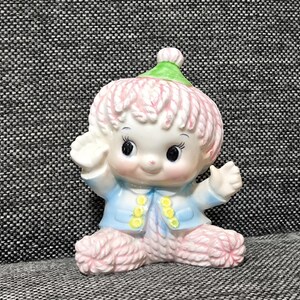 May include: A ceramic figurine of a seated character with pink textured hair, wearing a green pointed hat, a light blue jacket with yellow buttons, and pink textured legs. The character has large black eyes, a small nose, and raised arms.