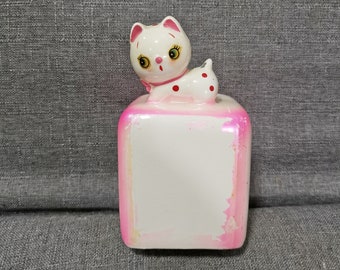 May include: A white ceramic cat figurine with pink polka dots and green eyes sits on top of a pink and white ceramic cube.