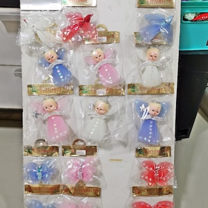 以下が含まれることがあります： クリスマスオーナメントのディスプレイ。天使のフィギュアと蝶の装飾が含まれています。天使は、キラキラ光る翼の白い、ピンク、青のドレスを着ています。蝶は青、ピンク、赤の色合いで、キラキラしたアクセントが付いています。各オーナメントは個別に包装されています。
